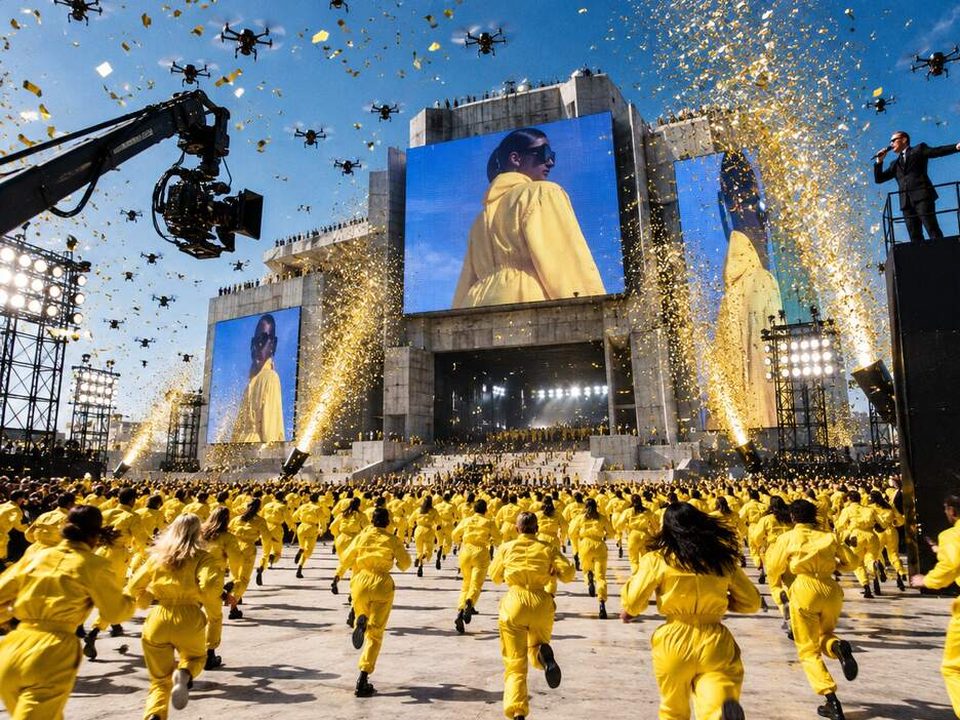 Hundreds of contestants in yellow jumpsuits sprint toward a building wrapped in giant screens, with confetti, drones, a camera crane and a presenter on a podium