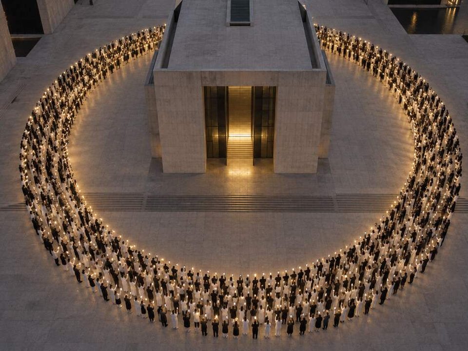 Aerial view at dusk: hundreds of people in white and black hold up glowing phones, forming a golden circle around a concrete building